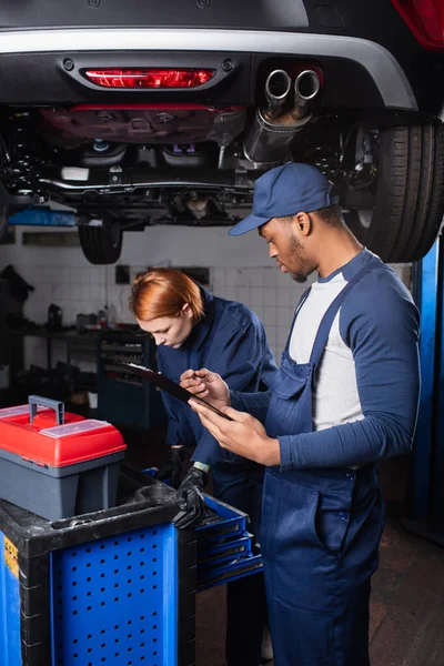 Afroamerikanischer Arbeiter hält Klemmbrett, während Kollege in der Nähe von Werkzeugkasten und Auto in der Garage arbeitet — Stockfoto