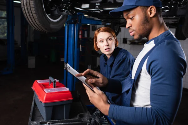 Mechanic talking to african american colleague with clipboard in car service — стоковое фото