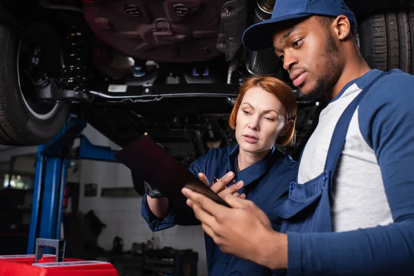 Interracial mechanics looking at clipboard near toolbox and car in garage — Fotografia de Stock