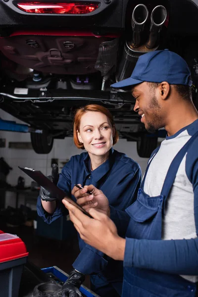 Mecánica multiétnica sonriente con portapapeles hablando cerca de caja de herramientas y coche en servicio — Stock Photo