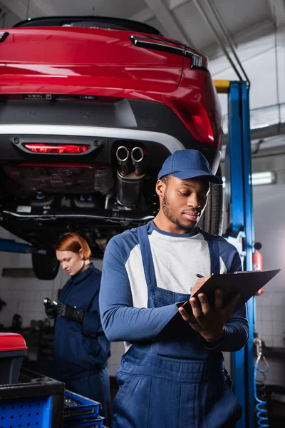 Afroamerikanischer Mechaniker in Uniform schreibt auf Klemmbrett neben Kollege und Auto in Garage — Stockfoto