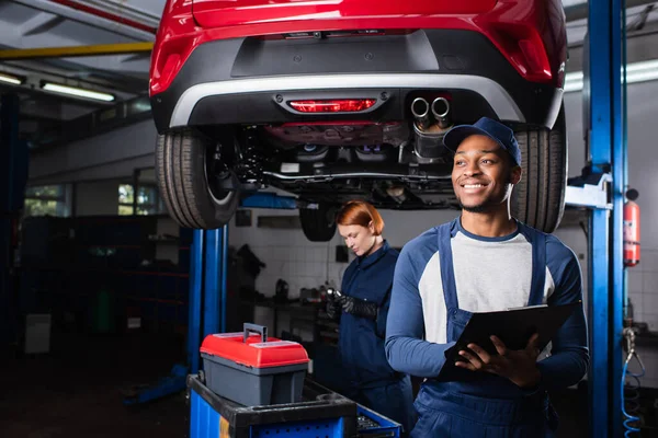 Young african american mechanic holding clipboard near colleague and car in service — Foto stock