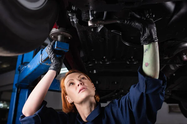 Pelirroja y mecánico tatuado trabajando bajo el coche en servicio - foto de stock