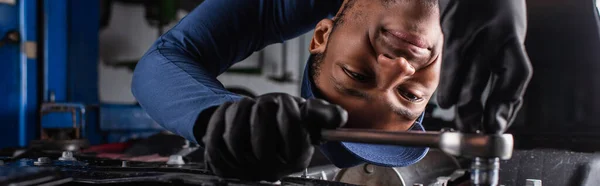 Trabajador afroamericano positivo usando la llave inglesa mientras que trabaja en servicio del coche, bandera - foto de stock