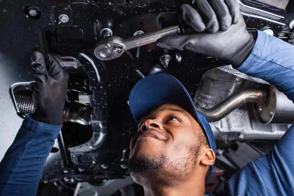 Vista de baixo ângulo do mecânico americano africano alegre que trabalha com chave inglesa sob o carro na garagem — Fotografia de Stock