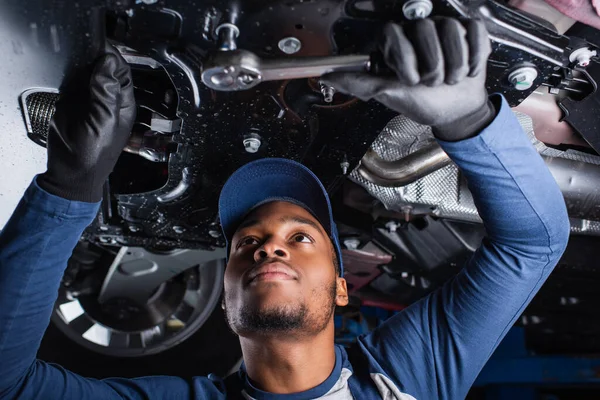 Joven afroamericano mecánico trabajando con herramienta debajo de coche en garaje - foto de stock