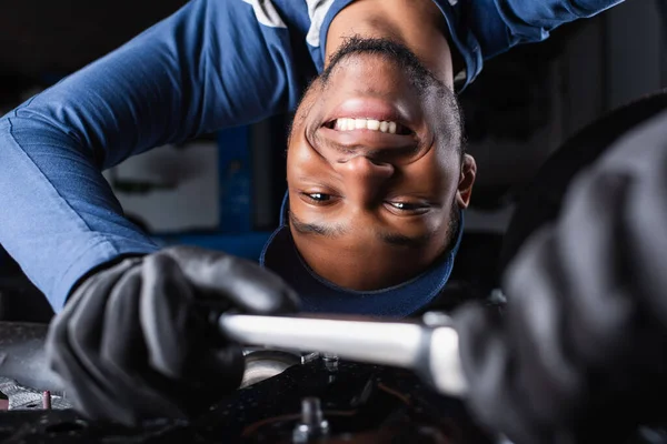 Heureux mécanicien afro-américain en gants travaillant sous voiture en service — Photo de stock