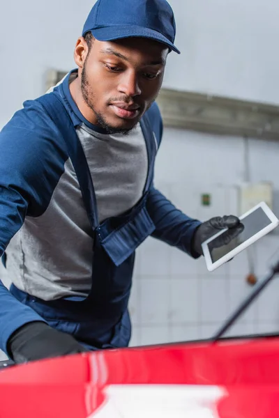Técnico afroamericano en uniforme sosteniendo la tableta digital con la pantalla en blanco cerca del coche borroso - foto de stock