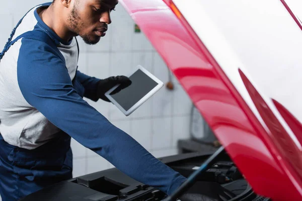 Joven afroamericano reparador sosteniendo tableta digital con pantalla en blanco mientras inspecciona el coche - foto de stock