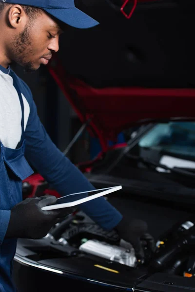 Joven afroamericano reparador sosteniendo tableta digital mientras que hace la inspección del coche borroso - foto de stock