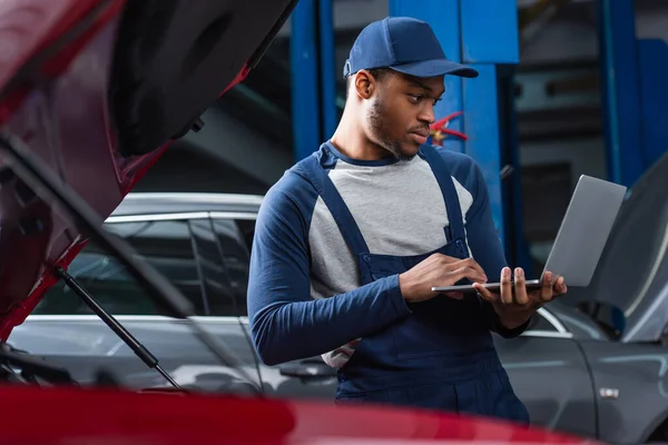 Junger afrikanisch-amerikanischer Polier benutzt Laptop in der Nähe verschwommener Autos in Werkstatt — Stockfoto