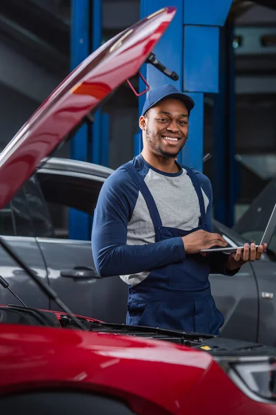 Sonriente afroamericano mecánico con portátil mirando a la cámara cerca de coches borrosos en el taller - foto de stock