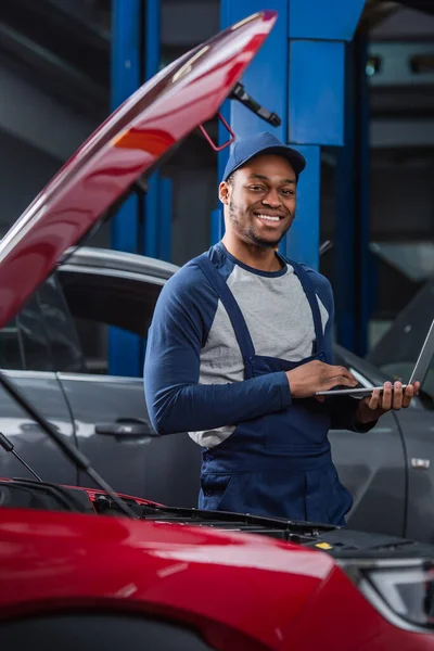 Meccanico afroamericano sorridente con computer portatile che guarda la macchina fotografica vicino alle auto sfocate in officina — Foto stock