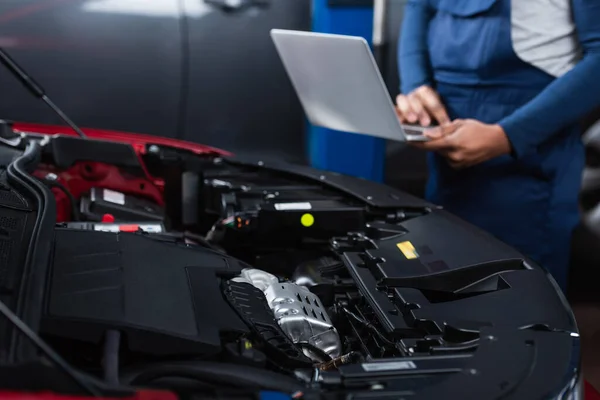 Partial view of blurred african american mechanic with laptop near car engine compartment — Stock Photo