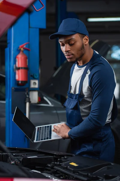 Africano americano reparador celebración de la computadora portátil con pantalla en blanco cerca del compartimiento del motor del coche - foto de stock