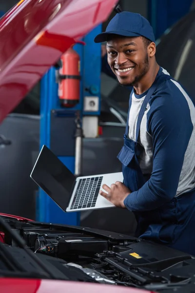Щасливий афро-американський механік, дивлячись на камеру, тримаючи ноутбук з порожнім екраном біля відсіку двигуна автомобіля — стокове фото