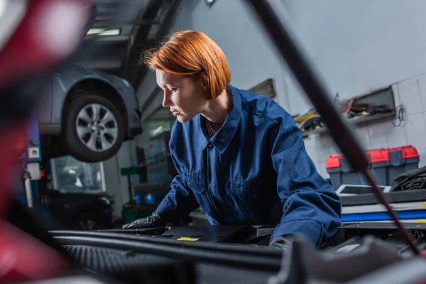 Forewoman in uniform inspecting blurred car with open hood in workshop — Foto stock