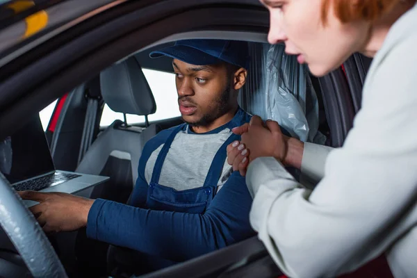 Afroamericano capataz apuntando a la computadora portátil mientras está sentado en el coche de un cliente borroso - foto de stock