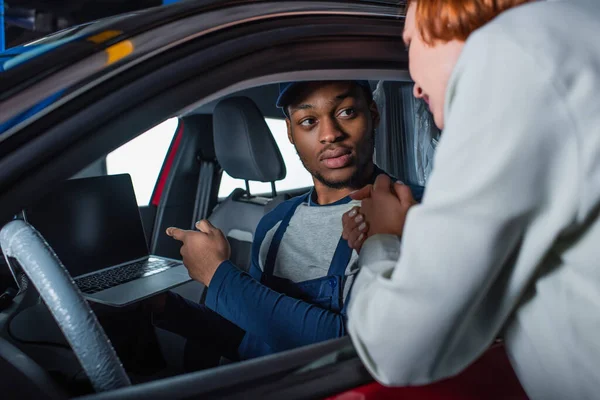 Técnico afroamericano apuntando a la computadora portátil mientras está sentado en el coche cerca del cliente borroso - foto de stock