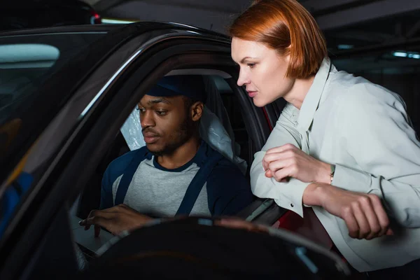 African american mechanic with laptop inspecting car near customer in workshop — Stock Photo