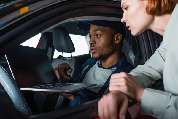 Afroamericano capataz apuntando a la computadora portátil mientras que hace diagnósticos de coche cerca del cliente - foto de stock