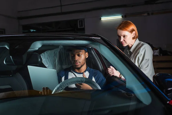 African american mechanic sitting in car with laptop near serious customer — Stock Photo
