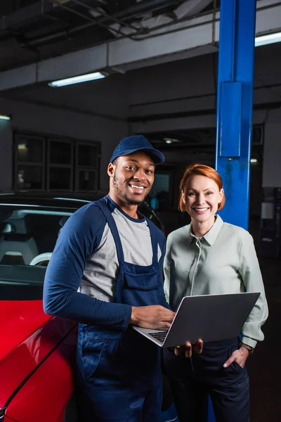 Happy client and african american mechanic with laptop smiling near car in workshop — Fotografia de Stock