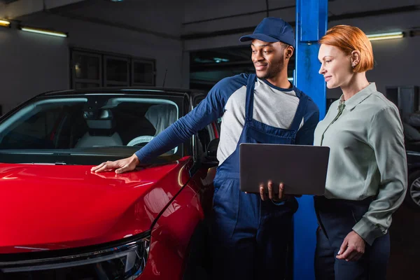 Jovem técnico americano africano com laptop de pé perto do carro e cliente satisfeito na estação de serviço — Fotografia de Stock