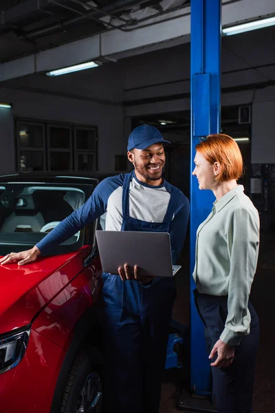 Contremaître afro-américain positif avec ordinateur portable parlant à un client souriant près de la voiture en atelier — Photo de stock