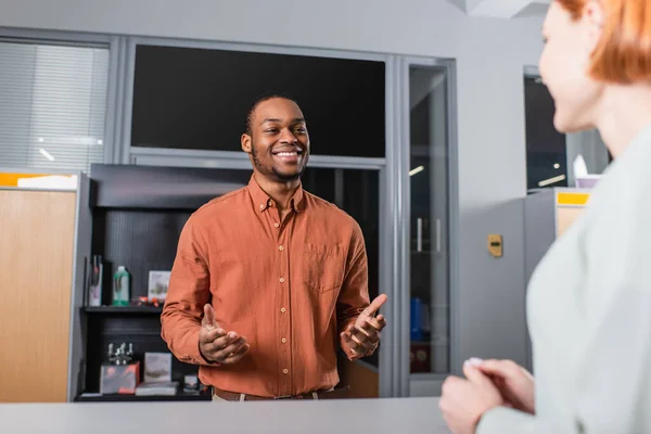 Cheerful african american car dealer talking to blurred woman near reception — Stock Photo