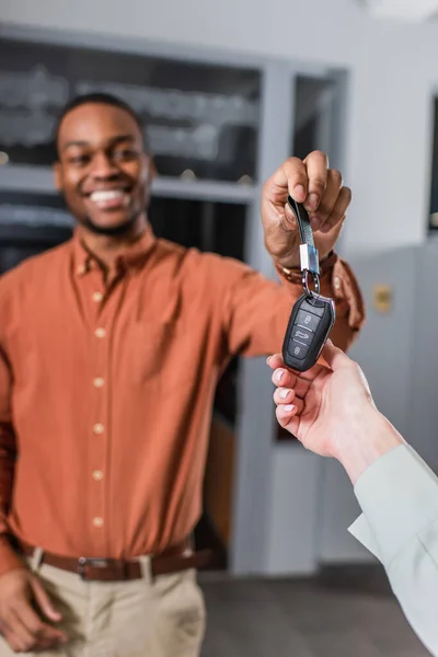 Concessionário de carros afro-americano borrado sorrindo, dando a chave para o cliente — Fotografia de Stock