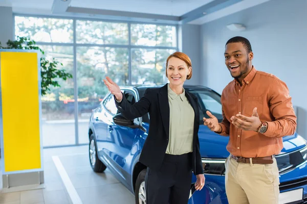 Smiling car dealer pointing with hand near amazed african american client — Stock Photo