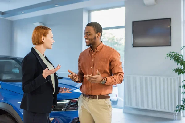 Smiling african american man talking to car dealer near auto in showroom — Foto stock