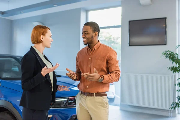 Sonriente afroamericano hombre hablando con concesionario de coches cerca de auto en sala de exposición - foto de stock