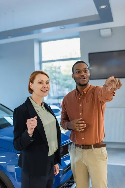 Sonriente afroamericano hombre mirando hacia otro lado y señalando con el dedo cerca de concesionario de coches - foto de stock