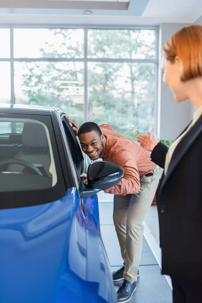 Blurred car dealer pointing with hand near happy african american man looking inside car in showroom — Stock Photo