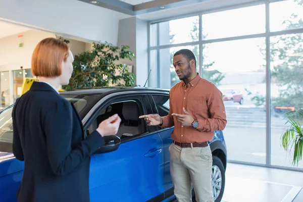 African american man pointing with hands at car while looking at blurred car dealer — Foto stock