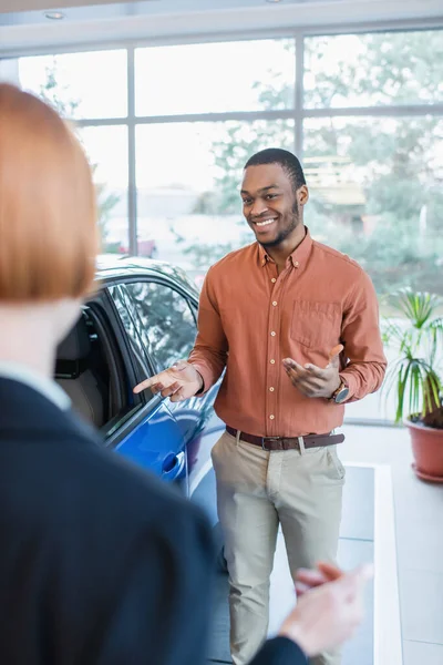 Hombre afroamericano complacido apuntando a auto cerca de distribuidor de coche borroso — Stock Photo