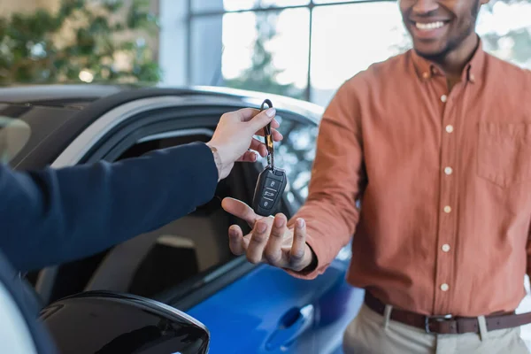 Обрізаний вид дилера автомобіля, що дає ключ афроамериканському клієнту, посміхаючись на розмитому фоні — стокове фото