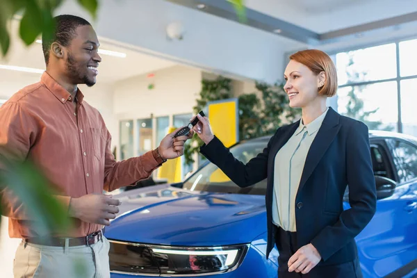 Concessionnaire de voiture souriant donnant la clé à l'homme afro-américain joyeux dans le showroom, au premier plan flou — Photo de stock