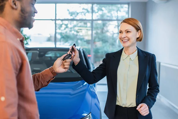 Happy car dealer giving key to blurred african american client near car in showroom — Stock Photo