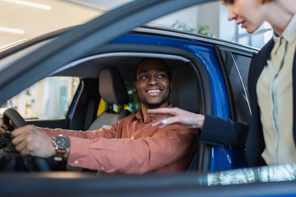 Blurred car dealer pointing with hand near happy african american owner sitting in car — Stock Photo