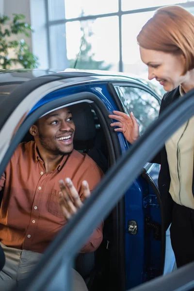 Happy african american man talking to blurred car dealer while sitting in auto — Stock Photo