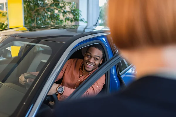 Cheerful african american man looking at blurred car dealer while sitting in car — Stockfoto