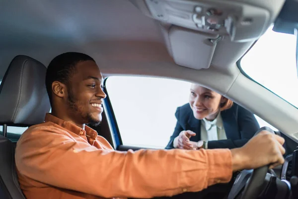 Concessionnaire de voiture floue pointant avec la main près de l'homme afro-américain joyeux assis dans la voiture — Photo de stock