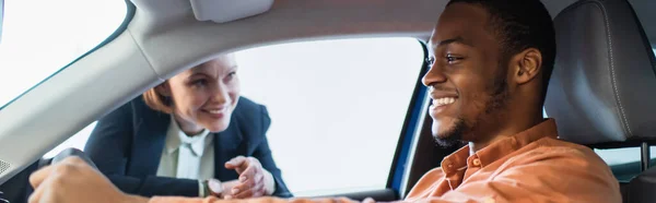 Hombre afroamericano feliz sentado en auto cerca de distribuidor de coche borroso, pancarta - foto de stock