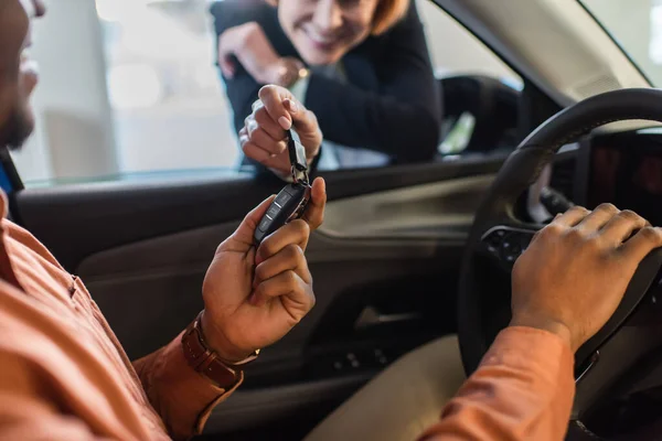 Partial view of blurred car dealer giving key to african american client sitting in car — стоковое фото