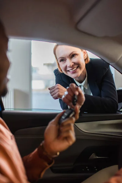 Negociante de carro alegre dando a chave para o comprador americano africano borrado sentado no carro — Fotografia de Stock