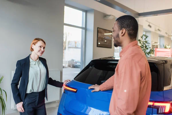 Concesionario de coches felices y difuminado cliente afroamericano de pie cerca de coche en sala de exposición - foto de stock