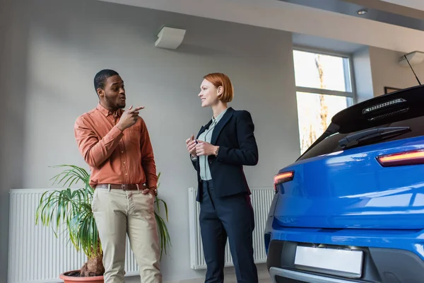 Homme afro-américain étonné pointant avec le doigt à l'auto près souriant concessionnaire de voitures — Photo de stock